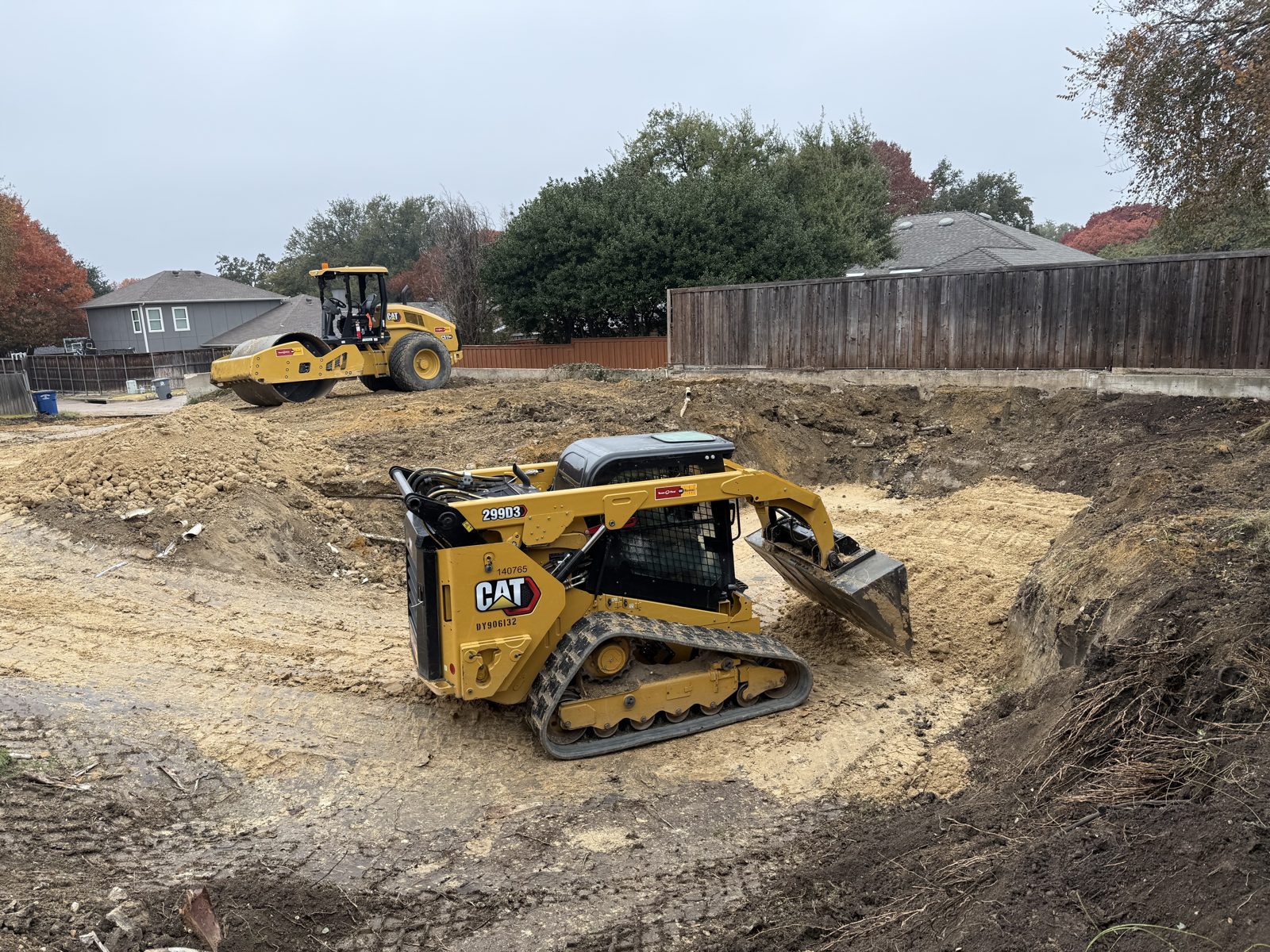 Skid steer and roller working an excavated cut in a fenced residential backyard